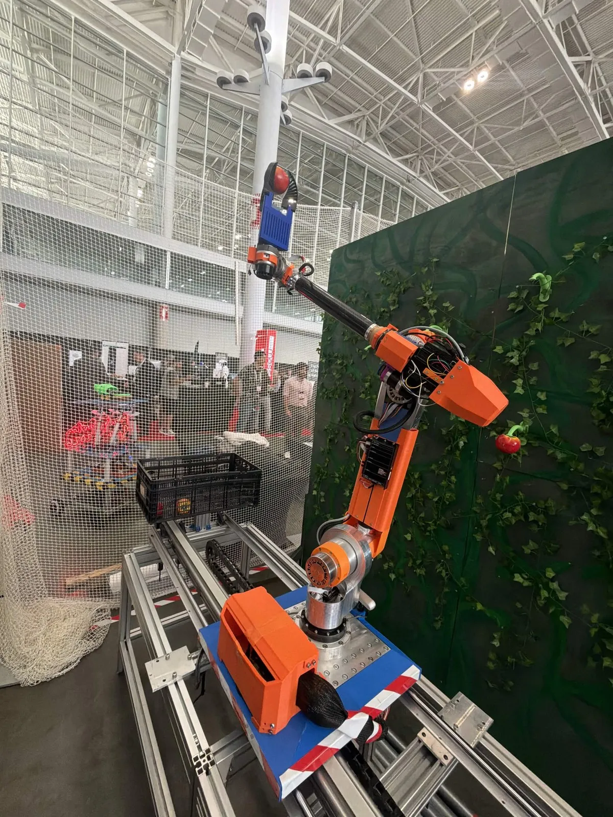 Robot arm on a rail holds a red tomato in its gripper inside a netted arena at an expo hall, with a collection crate nearby.