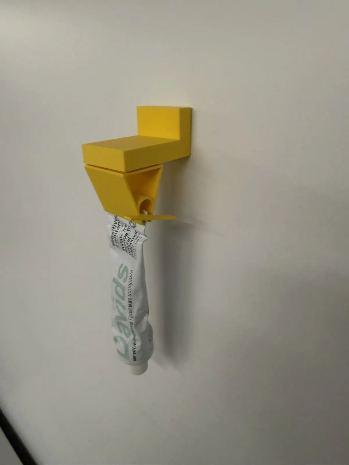 Straight-on view of a yellow 3D-printed dispenser bracket holding a partly squeezed toothpaste tube on a white wall.