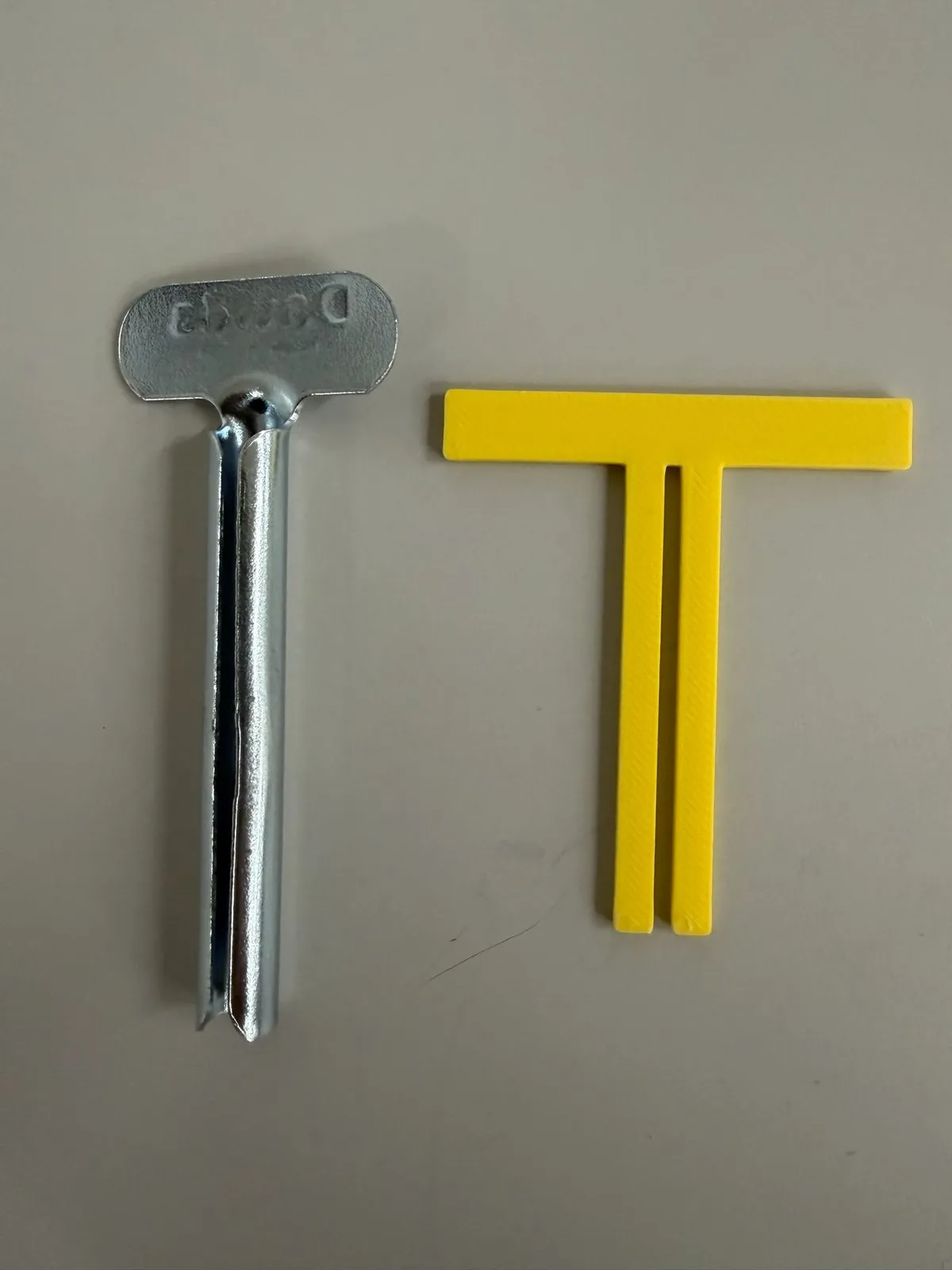 Commercial Davids stamped-steel tube key lying next to a yellow 3D-printed T-shaped key prototype on a gray surface.