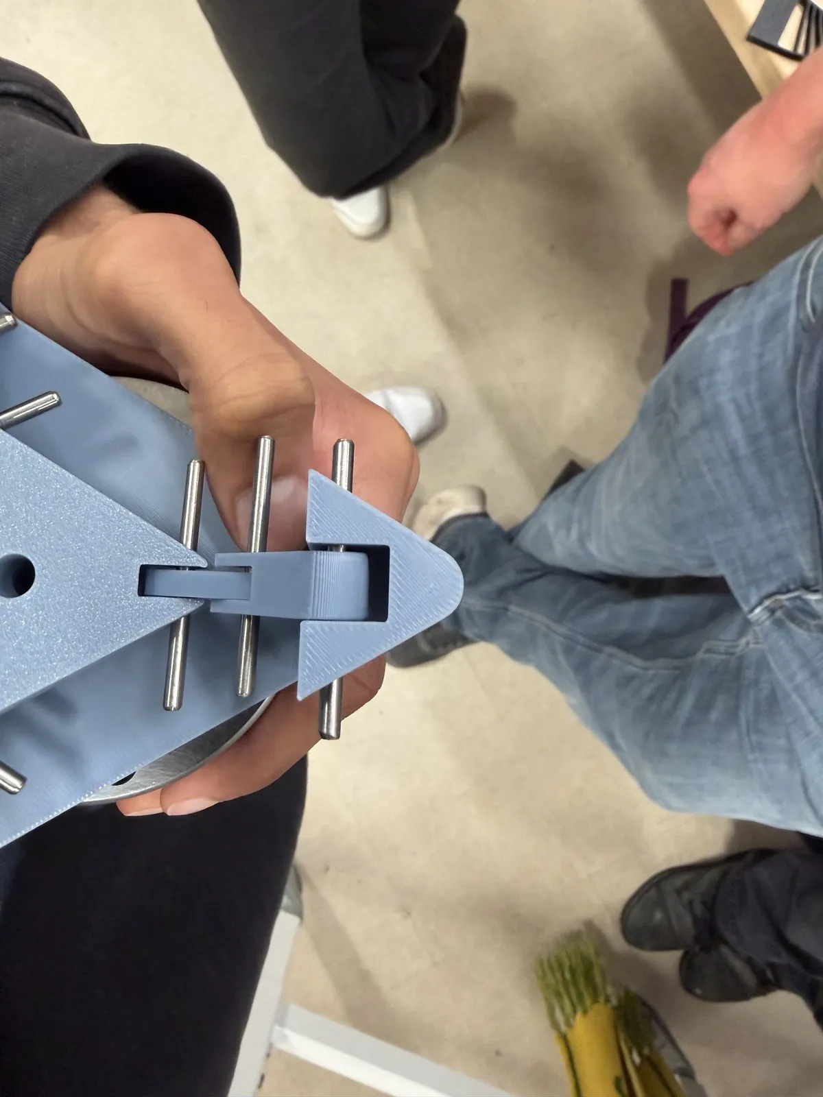 Hand holding a gray-blue 3D-printed corner piece with steel dowel pins and a printed sliding latch, expo floor below.