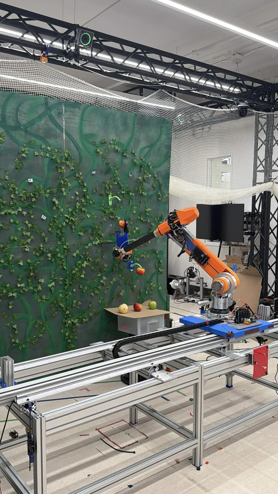 Robot arm on an aluminum-rail gantry reaching toward a red tomato on a green vine-covered wall in a netted test cell.